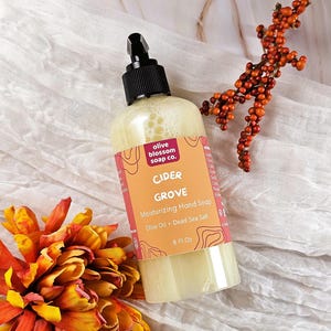 May include: A clear plastic bottle of Cider Grove moisturizing hand soap with a black pump dispenser. The label is orange with red accents, featuring the text olive blossom soap co., Cider Grove, Moisturizing Hand Soap, Olive Oil + Dead Sea Salt, 8 Fl Oz.