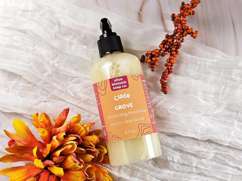 May include: A clear plastic bottle of Cider Grove moisturizing hand soap with a black pump dispenser. The label is orange with red accents, featuring the text olive blossom soap co., Cider Grove, Moisturizing Hand Soap, Olive Oil + Dead Sea Salt, 8 Fl Oz.
