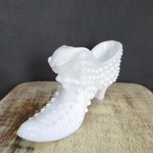 May include: A white milk glass shoe shaped trinket dish. The shoe has a pointed toe and a high heel. The surface of the shoe is covered in small, raised dots.