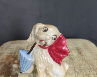 May include: A white ceramic figurine of a dog holding a blue umbrella and wearing a red bow. The dog has large, black eyes and a playful expression.