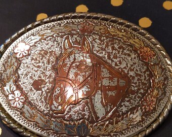 May include: Oval-shaped silver and copper-toned belt buckle featuring a horse head design. The buckle has a decorative floral and leaf border. The horse head is in the center of the buckle, with a distressed finish.