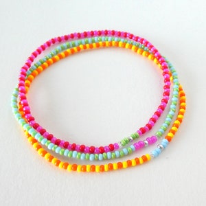 Puede incluir: Un brazalete de cuentas multicolores con tres hileras de cuentas en naranja, amarillo, verde, rosa y azul claro.