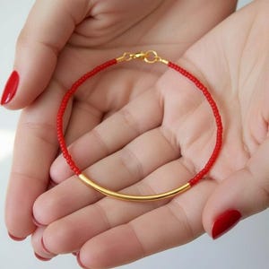 May include: A red beaded bracelet with a gold-colored curved bar. The bracelet is held in open hands with red nail polish. The bracelet is a delicate piece of jewelry.