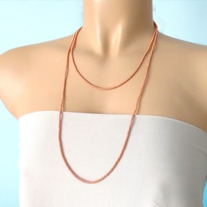 Puede incluir: Un collar largo y delicado de cuentas de color cobre drapeado sobre un maniquí. El collar está compuesto por pequeñas cuentas, creando un hilo continuo. Las cuentas tienen un brillo metálico, que refleja la luz. El collar es un accesorio sencillo pero elegante.