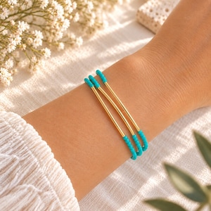 Könnte beinhalten: Ein dreireihiges Armband mit drei goldfarbenen Metallstäben, die sich mit Abschnitten aus kleinen, leuchtend türkisfarbenen Perlen abwechseln. Das Design besteht aus drei parallelen Linien, die das Handgelenk umschließen und metallische Elemente mit farbigen Perlenabschnitten für einen geschichteten Look kombinieren.