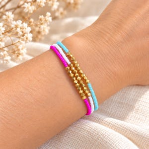 Può includere: Un set di tre braccialetti di perline con perline di conteria in rosa vivace, bianco e azzurro. Ogni braccialetto include una sezione centrale di piccole perline metalliche dorate sfaccettate che aggiungono una consistenza riflettente al design.
