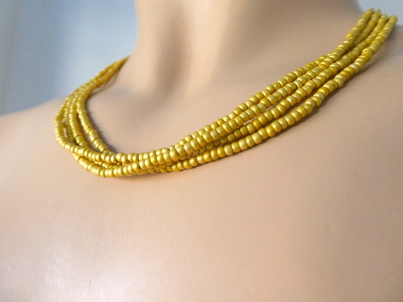 May include: A gold-colored beaded necklace with multiple strands. The necklace is made of small, round beads and is worn on a mannequin.