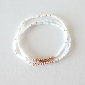May include: Three delicate beaded bracelets. The bracelets feature white seed beads with small silver-colored accents. A section of rose gold-colored faceted beads adds a touch of color to each bracelet. The bracelets are displayed on a white surface.