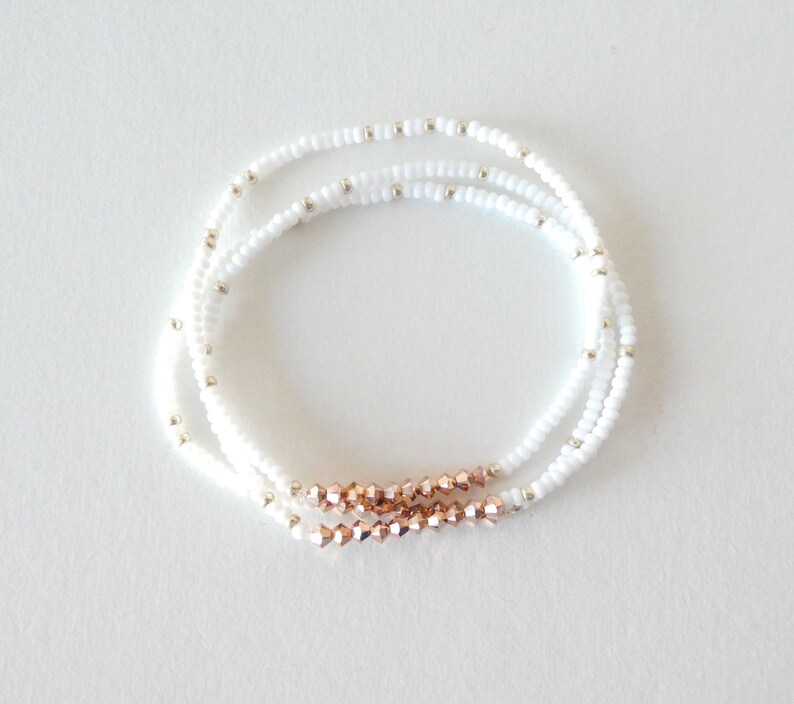 May include: Three delicate beaded bracelets. The bracelets feature white seed beads with small silver-colored accents. A section of rose gold-colored faceted beads adds a touch of color to each bracelet. The bracelets are displayed on a white surface.