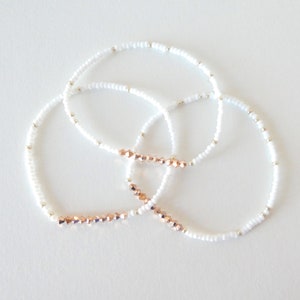 May include: Three delicate bracelets. Each bracelet features small white beads and silver accents. One section of each bracelet is adorned with rose gold-colored faceted beads. The bracelets are arranged in a circular pattern against a white background.