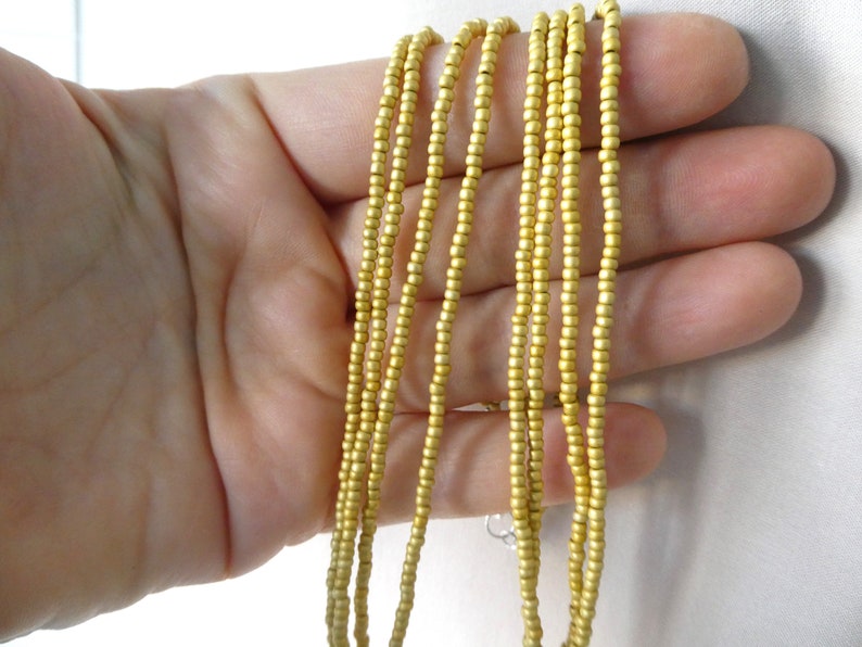 May include: A strand of gold-colored beads, made of small, round beads, is shown in a hand. The beads are strung together to form a long necklace.