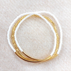 May include: Three beaded bracelets. Two bracelets are white with small beads, and one is gold with small beads. The bracelets have gold curved bars at the ends. The bracelets are stacked on top of each other.