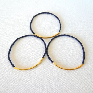 May include: Three navy blue beaded bracelets with a gold bar accent. The bracelets are stacked on top of each other.