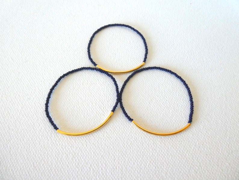 May include: Three navy blue beaded bracelets with a gold bar accent. The bracelets are stacked on top of each other.