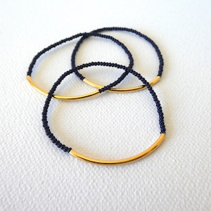 May include: Three gold and black beaded bracelets with a gold bar accent. The bracelets are stacked on top of each other.