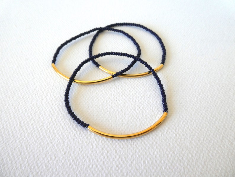 May include: Three gold and black beaded bracelets with a gold bar accent. The bracelets are stacked on top of each other.