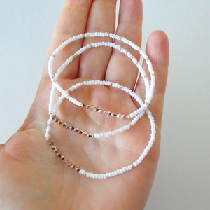 May include: Three delicate bracelets with white seed beads and rose gold faceted beads. The bracelets are strung on a clear elastic cord and are displayed on a hand.