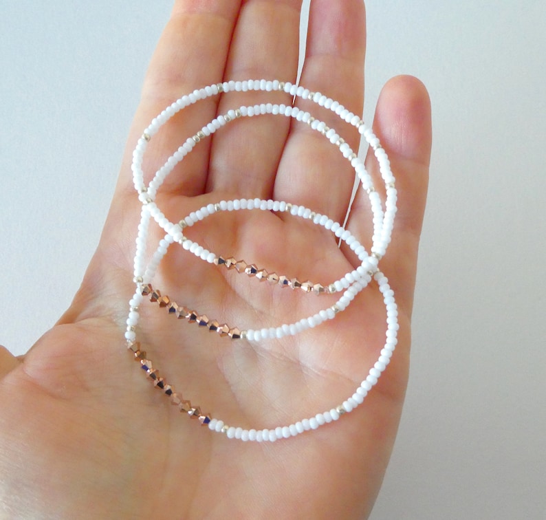 May include: Three delicate bracelets with white seed beads and rose gold faceted beads. The bracelets are strung on a clear elastic cord and are displayed on a hand.