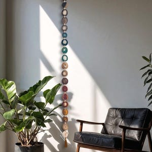 May include: A vertical wall hanging featuring a long string of circular, multi-colored agate slices in shades of blue, green, brown, and purple. The stones are connected by small white beads, and the piece terminates with a brown tassel at the bottom.