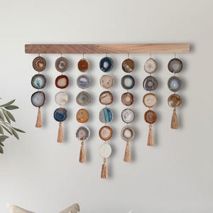 May include: A wall hanging featuring a horizontal wooden bar with seven vertical strands of circular agate slices in various shades of blue, brown, gray, and white. Each strand ends with a small, light brown tassel hanging from the bottom stone.