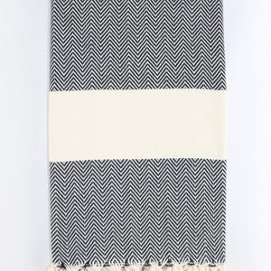 May include: A folded black and white herringbone patterned towel with a cream-colored stripe across the center. The towel has cream-colored fringe along the bottom edge. The towel is made of a soft, absorbent material.