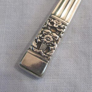 Puede incluir: Primer plano de un mango de utensilio plateado con detalles florales. El mango tiene forma rectangular con un patrón floral detallado en el centro y líneas verticales en la parte superior. La parte inferior del mango es lisa y rectangular.