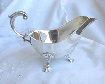 Könnte beinhalten: Eine polierte silberfarbene Sauciere mit geschwungenem Griff, breitem Ausguss und drei dekorativen Füßen, die oben jeweils mit einem muschelartigen Muster verziert sind. Die metallische Oberfläche ist glatt und reflektierend gestaltet.