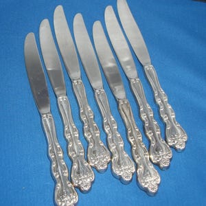 Può includere: Set di sette coltelli color argento con manici ornati e dettagliati. Le lame sono lisce e i manici presentano un design decorativo floreale. I coltelli sono disposti su una superficie blu, suggerendo un servizio da tavola o posate.