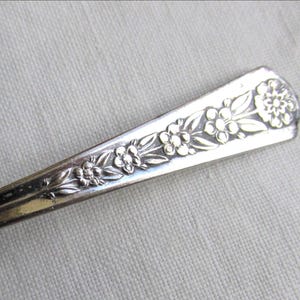 May include: A silver-toned metal spoon handle featuring an embossed floral pattern. The design consists of a series of small, five-petaled flowers and elongated leaves arranged in a row along the tapered, slightly curved handle of the utensil.
