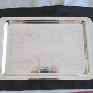 May include: A rectangular silver serving tray with ornate handles. The tray has a reflective surface with a subtle floral pattern. The handles are curved and feature a decorative edge. The tray is likely used for serving food or drinks.