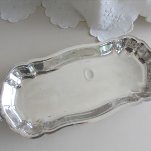 May include: A rectangular silver-toned serving tray with a scalloped edge and two ornate, curved handles on opposite ends. The tray features a polished, reflective surface with a smooth, flat base and raised, decorative sides for containment.