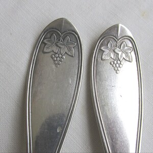 Puede incluir: Primer plano de dos cucharas plateadas. Cada cuchara presenta un diseño detallado de uvas y hojas en la parte superior del mango. Las cucharas tienen un aspecto clásico y elegante, perfecto para una colección vintage o antigua.