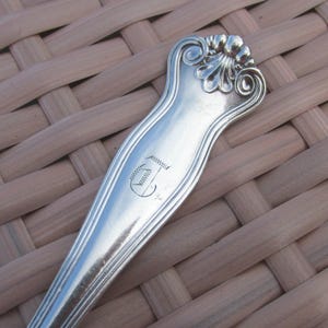 May include: Close-up of a silver spoon with an ornate handle. The handle features a floral design at the top and a monogrammed letter "J" in the center. The spoon is set against a woven background.