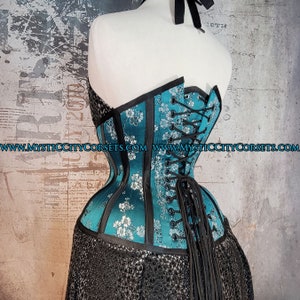 May include: A teal blue corset with black trim and silver floral embroidery. The corset has a lace-up back with black laces and a long tail. The corset is made of satin and has a smooth, shiny finish. The website address "www.mysticcitycorsets.com" is printed on the corset.
