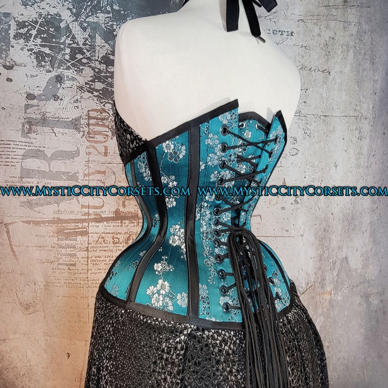 May include: A teal blue corset with black trim and silver floral embroidery. The corset has a lace-up back with black laces and a long tail. The corset is made of satin and has a smooth, shiny finish. The website address "www.mysticcitycorsets.com" is printed on the corset.
