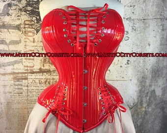Op de afbeelding: Een rood patentleren korset met een hartvormige halslijn en rode lintjes om te strikken. Het korset heeft een busk-sluiting aan de voorkant en is gemaakt van een glanzend, glad materiaal. Het websiteadres "www.mysticcitycorsets.com" is afgedrukt op de voorkant van het korset.