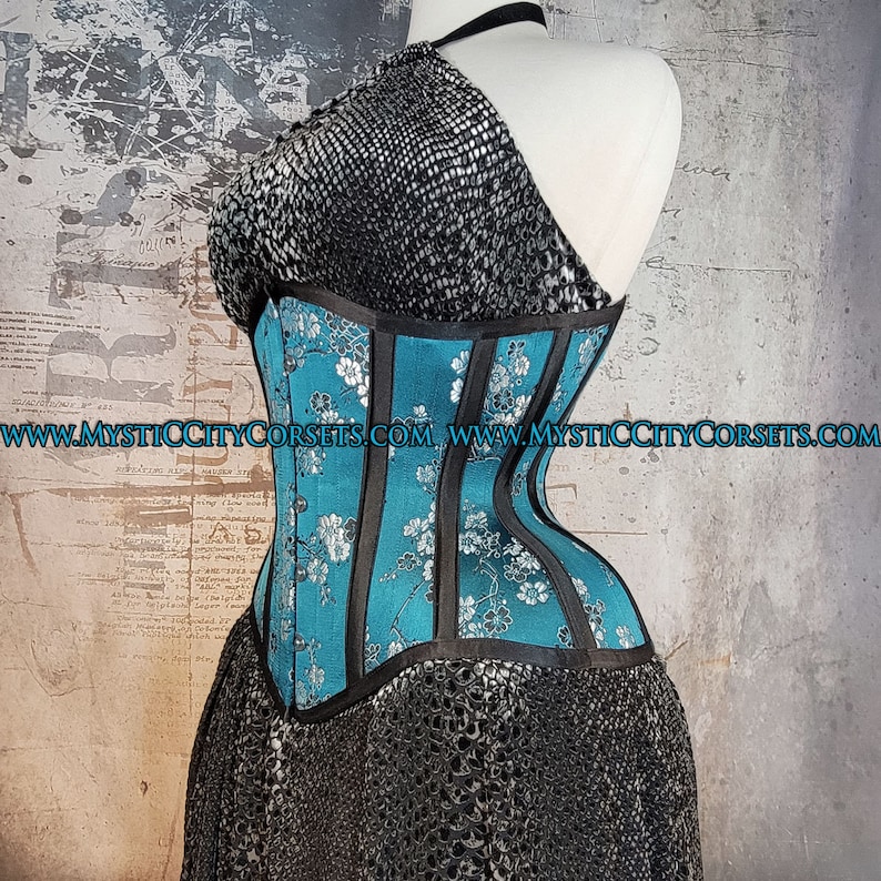 May include: A turquoise blue corset with black satin ribbon accents and a floral pattern. The corset is made of a smooth, silky fabric and has a boned structure. The corset is shown on a mannequin against a textured background.