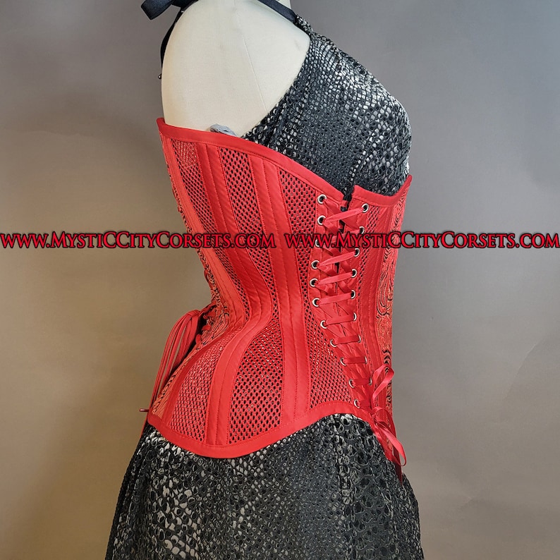 May include: A red corset with a mesh design and a lace-up back. The corset is made of a satin-like fabric and has a boned structure. The corset is shown on a mannequin and has the website address "www.mysticcitycorsets.com" printed on the bottom.