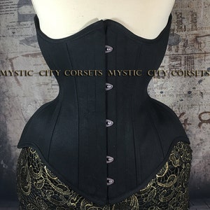 May include: A black corset with a structured design, featuring a row of buttons down the front. The corset is displayed on a mannequin. The bottom edge is trimmed with gold lace. The text "MYSTIC CITY CORSETS" is visible.
