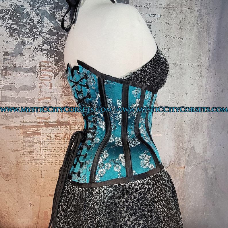May include: A turquoise blue corset with black trim and white floral embroidery. The corset has a lace-up back and is made of satin.