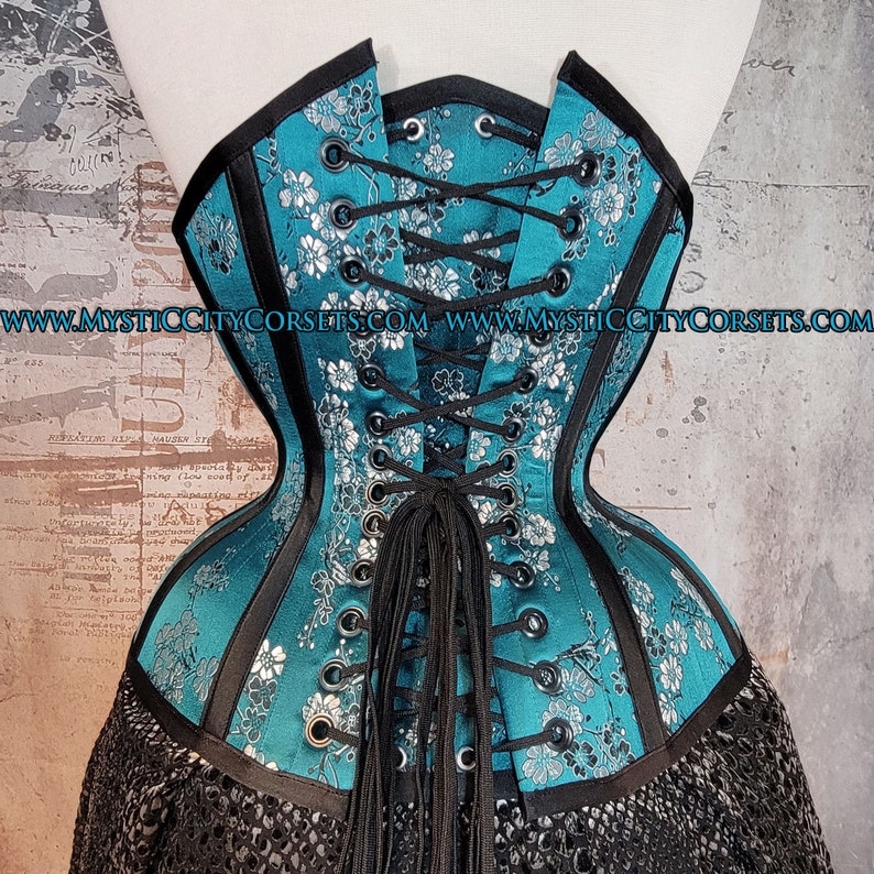 May include: A teal blue corset with black trim and silver floral embroidery. The corset laces up the back with black laces and has a black tassel.