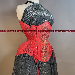 May include: Red corset with a black and gold floral design and a mesh panel. The corset has a lace-up back and is made of a satin-like fabric. The website address "www.mysticcitycorsets.com" is printed on the corset.