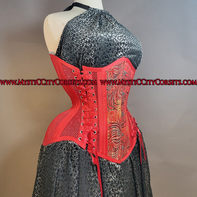 May include: Red corset with a black and gold floral design and a mesh panel. The corset has a lace-up back and is made of a satin-like fabric. The website address "www.mysticcitycorsets.com" is printed on the corset.