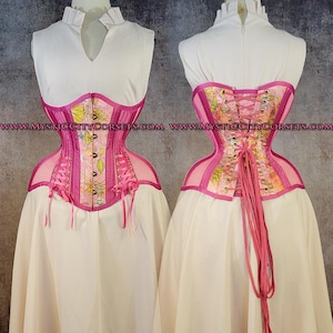 May include: A pink and floral patterned corset featuring a front busk closure with metal clasps and a back lace-up system with long pink ribbons. The garment displays the text WWW.MYSTICCITYCORSETS.COM and is worn over a white sleeveless top.