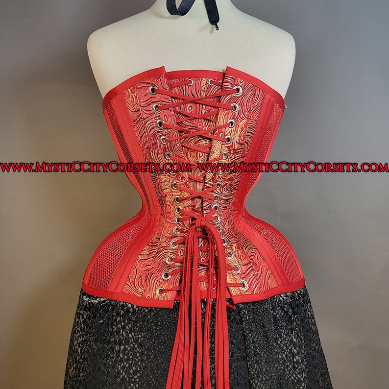 May include: A red corset with a gold and red patterned design and a lace-up back. The corset is made of a stretchy fabric and has a mesh panel on the sides. The corset is tied with red laces and has a long red tassel.