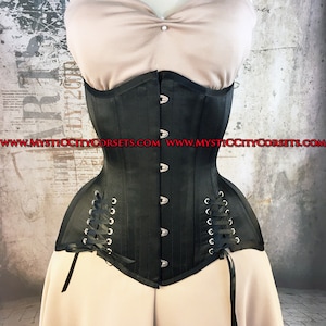 Pode incluir: Um corpete de cetim preto com fecho de busk e atadura nas costas. O corpete tem um acabamento liso e elegante e apresenta ilhós e ganchos de metal prateados. O corpete é mostrado em um manequim com um fundo bege.