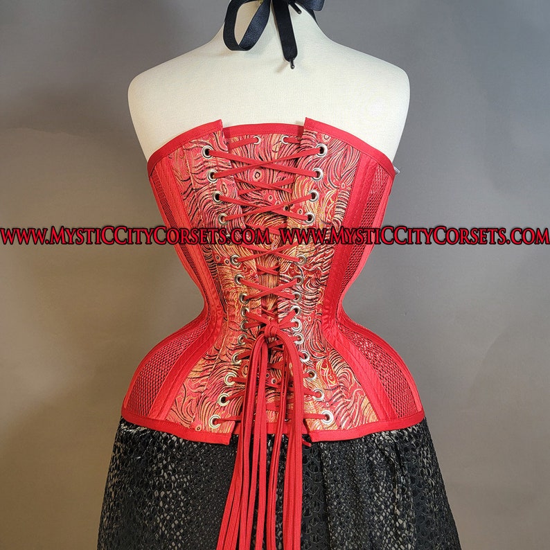 May include: A red corset with a floral pattern and a black ribbon. The corset is laced up the back and has a mesh panel on the sides. The corset is on a mannequin and the text "www.mysticcitycorsets.com" is visible on the bottom of the image.