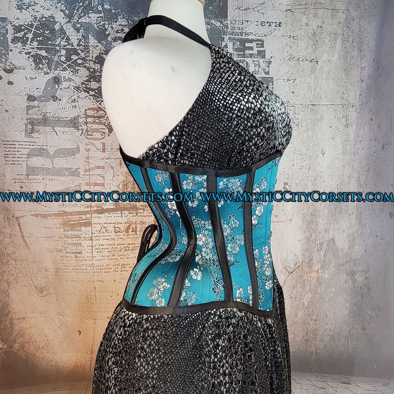 May include: A turquoise blue corset with black satin ribbon accents and a floral pattern. The corset is worn over a black skirt with a textured pattern. The text "www.mysticcitycorsets.com" is visible on the image.