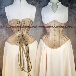 May include: Two beige corsets displayed on mannequins. The corsets have a structured design with vertical panels and lace-up backs. The fabric appears to be a satin-like material with mesh detailing. The mannequins are wearing white tops and skirts.