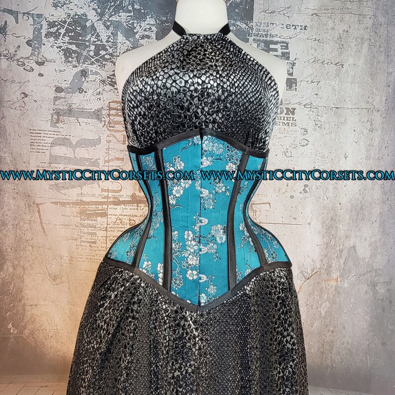 May include: A turquoise blue corset with black boning and floral embroidery. The corset has a halter top with a silver snakeskin pattern. The text "www.mysticcitycorsets.com" is visible on the bottom of the image.